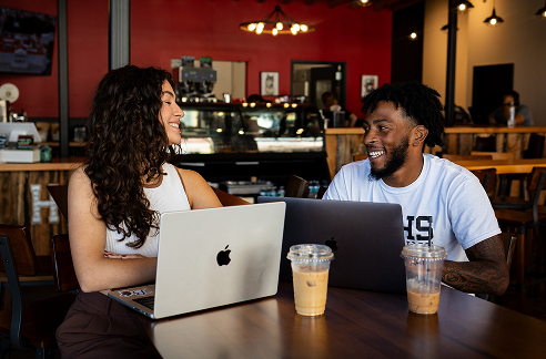 Two students having coffee sitting at a cafe with two laptops in front of them.
